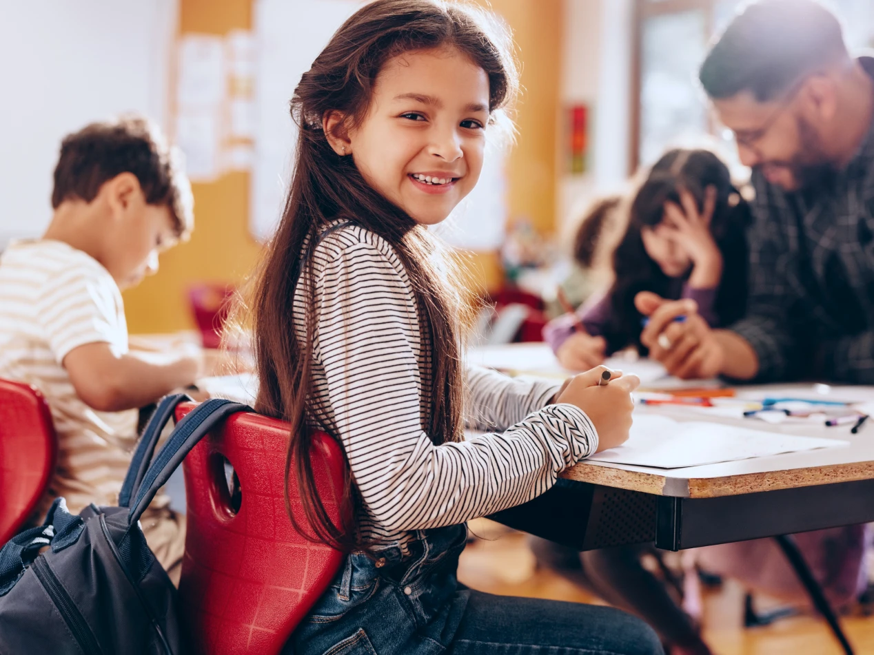 Smiling child sits at desk in a classroom environment.