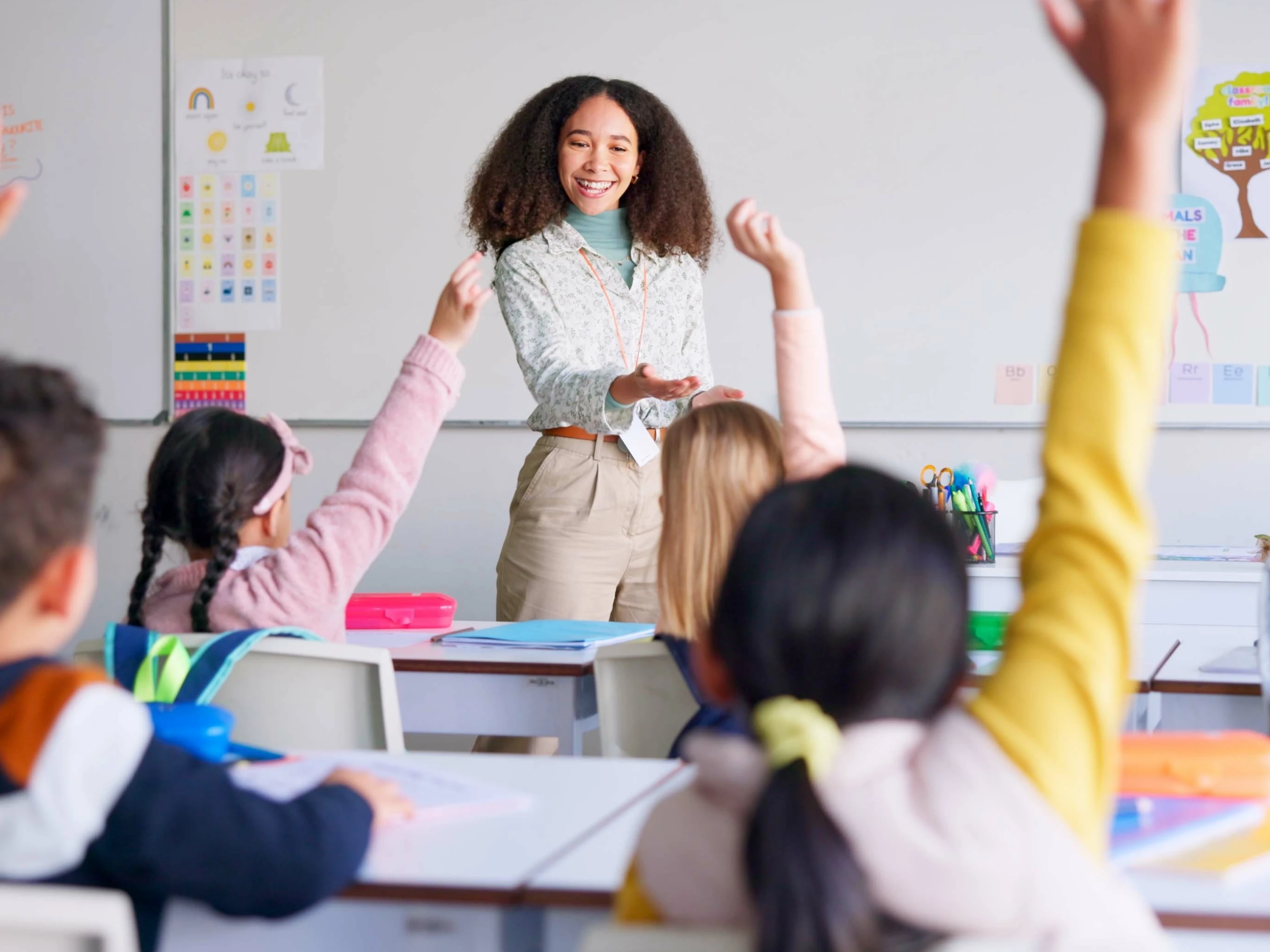 Teacher stands in front of classroom as students raise their hands.