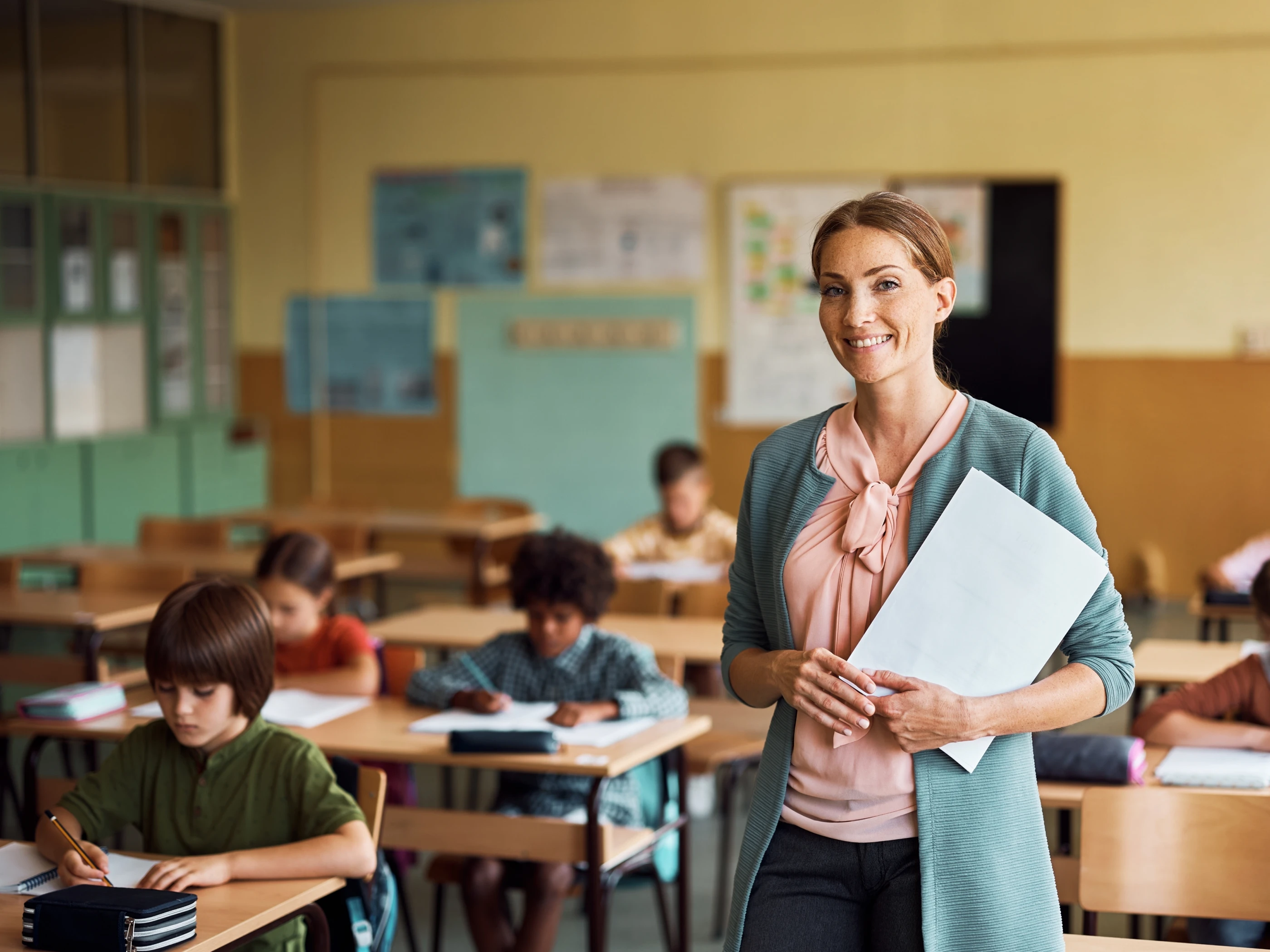 Happy school teacher stands holding folders in a classroom